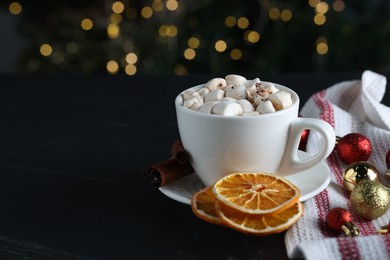 Photo of Tasty hot cocoa drink with marshmallows in cup, dry orange slices and baubles on black table. Space for text