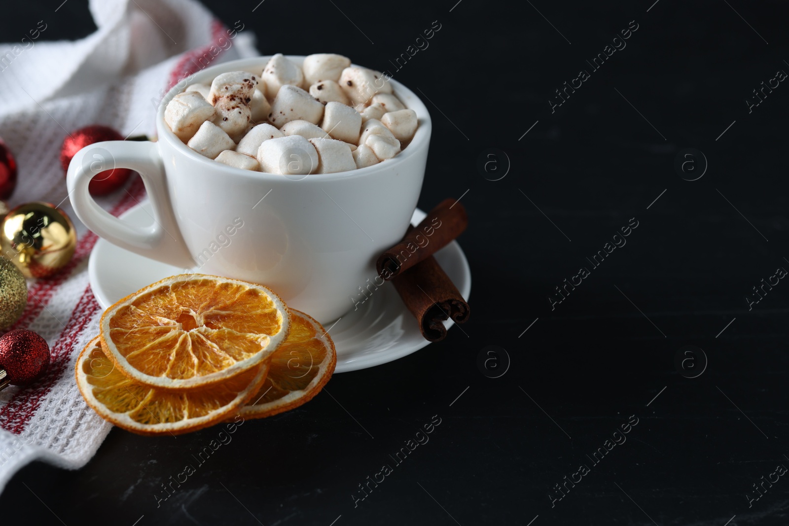 Photo of Tasty hot cocoa drink with marshmallows in cup, dry orange slices and baubles on black table. Space for text