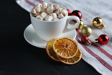 Photo of Tasty hot cocoa drink with marshmallows in cup, dry orange slices and baubles on black table