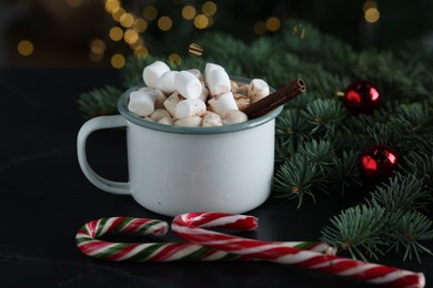 Photo of Tasty hot cocoa drink with marshmallows in mug, candy canes and fir branches on black table, closeup