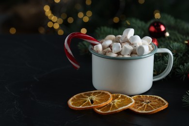 Photo of Tasty hot cocoa drink with marshmallows, candy cane in mug and dry orange slices on black table, closeup. Space for text