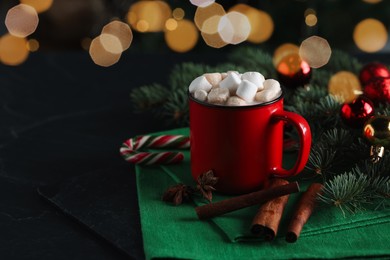 Photo of Tasty hot cocoa drink with marshmallows in mug, spices and fir branches on black table, closeup. Space for text