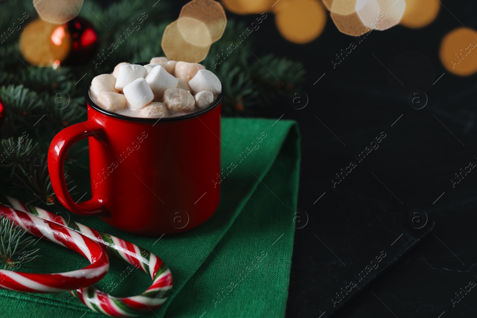 Photo of Tasty hot cocoa drink with marshmallows in mug, candy canes and fir branches on black table, closeup. Space for text