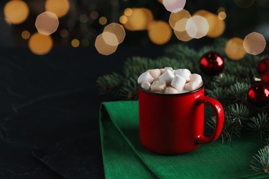 Photo of Tasty hot cocoa drink with marshmallows in mug, candy canes and fir branches on black table, closeup. Space for text