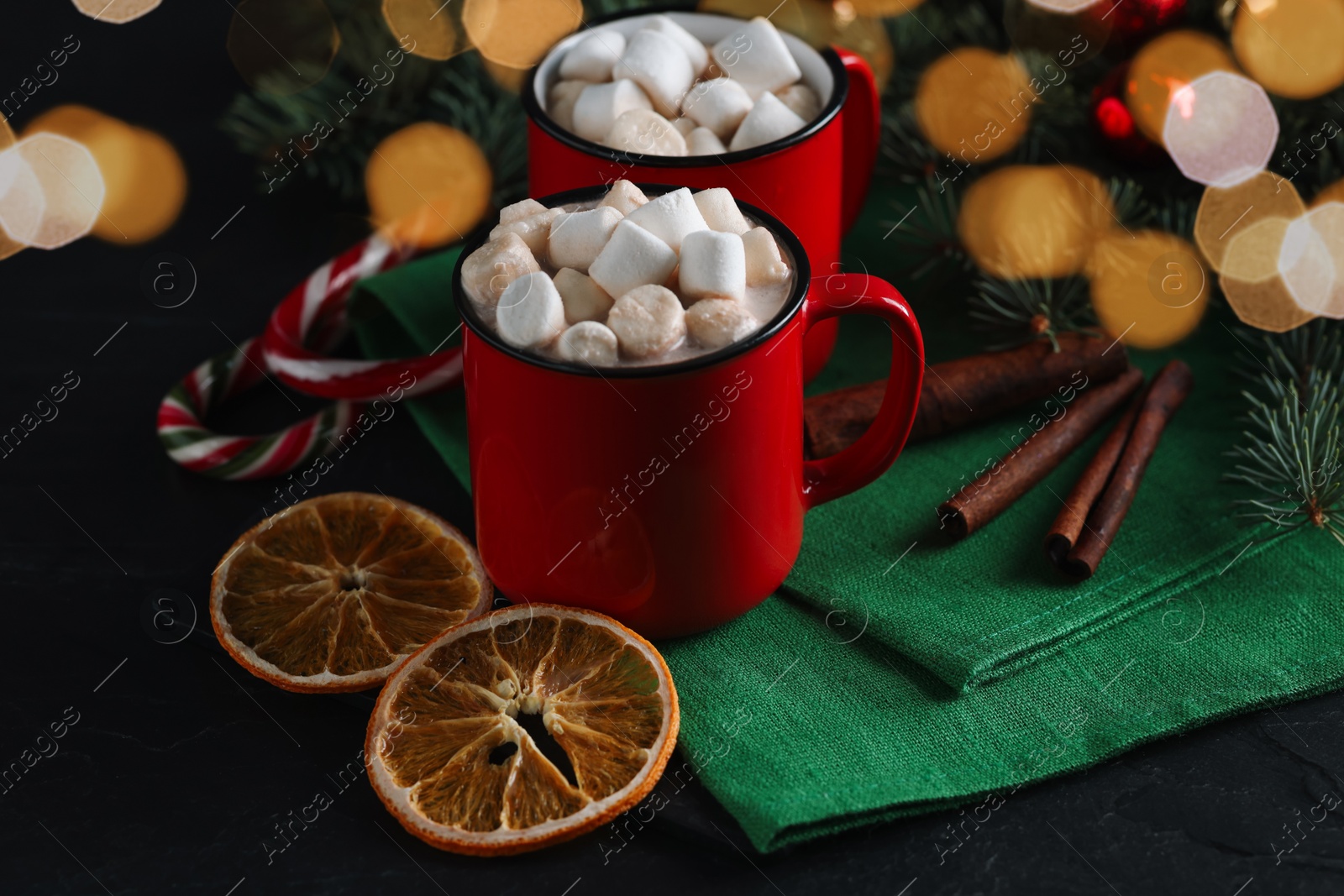 Photo of Tasty hot cocoa drinks with marshmallows in mugs and dry orange slices on black table, closeup