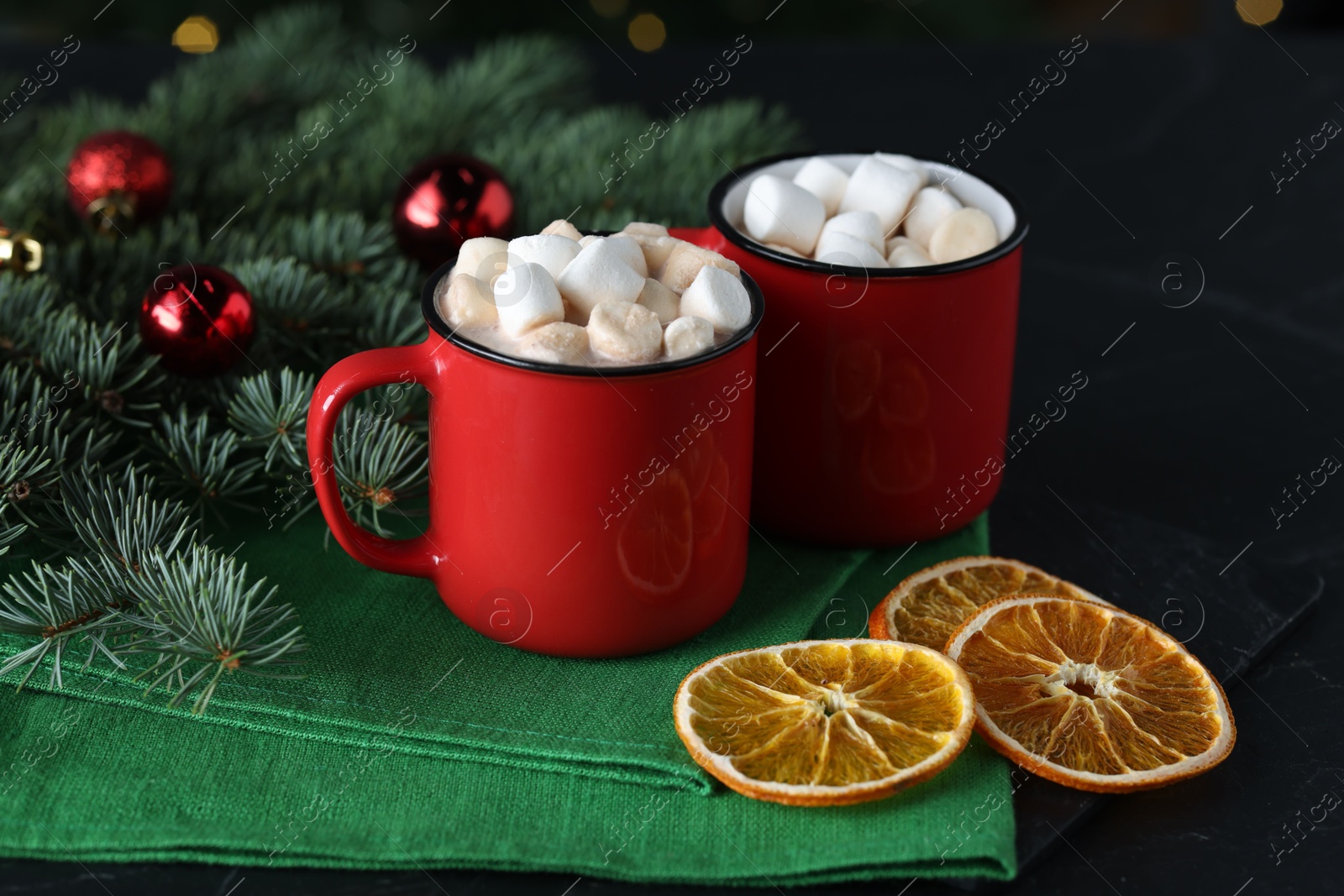 Photo of Tasty hot cocoa drinks with marshmallows in mugs, fir branches and dry orange slices on black table, closeup