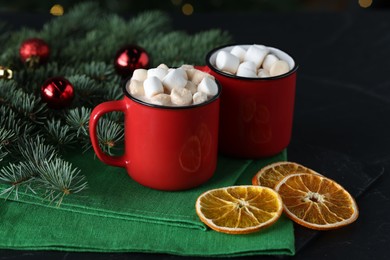 Photo of Tasty hot cocoa drinks with marshmallows in mugs, fir branches and dry orange slices on black table, closeup