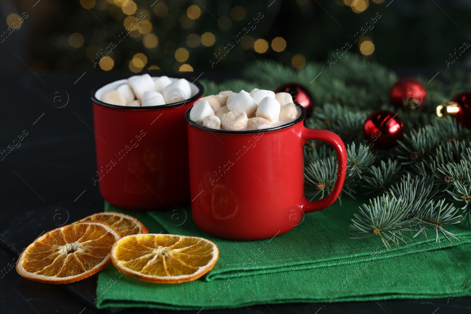 Photo of Tasty hot cocoa drinks with marshmallows in mugs, fir branches and dry orange slices on black table, closeup