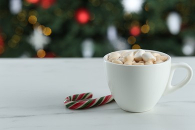 Photo of Tasty hot cocoa drink with marshmallows in cup and candy cane on white table against blurred lights. Space for text