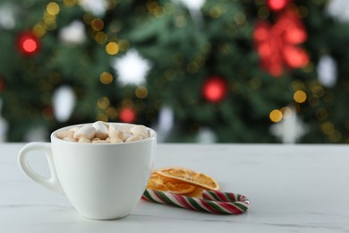 Photo of Tasty hot cocoa drink with marshmallows in cup, dry orange slices and candy cane on white table against blurred lights. Space for text