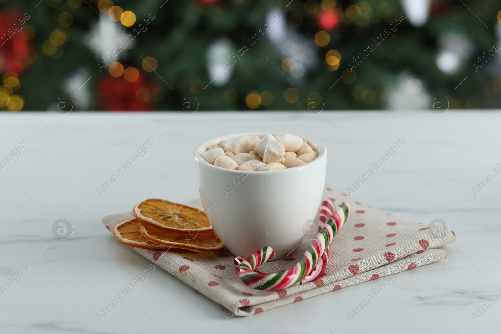 Photo of Tasty hot cocoa drink with marshmallows in cup, dry orange slices and candy canes on white table against blurred lights