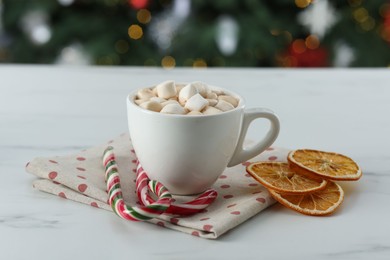 Photo of Tasty hot cocoa drink with marshmallows in cup, dry orange slices and candy canes on white table against blurred lights