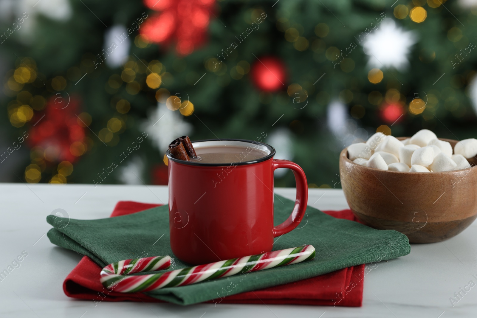 Photo of Tasty hot cocoa drink with cinnamon sticks in mug, candy cane and marshmallows on white table against blurred lights