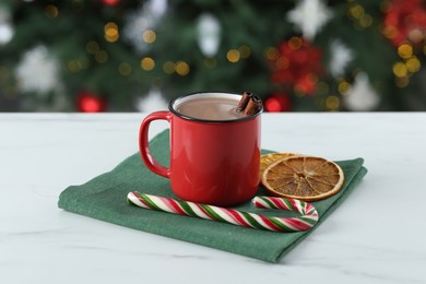 Photo of Tasty hot cocoa drink with cinnamon sticks in mug, candy cane and dry orange slices on white table against blurred lights