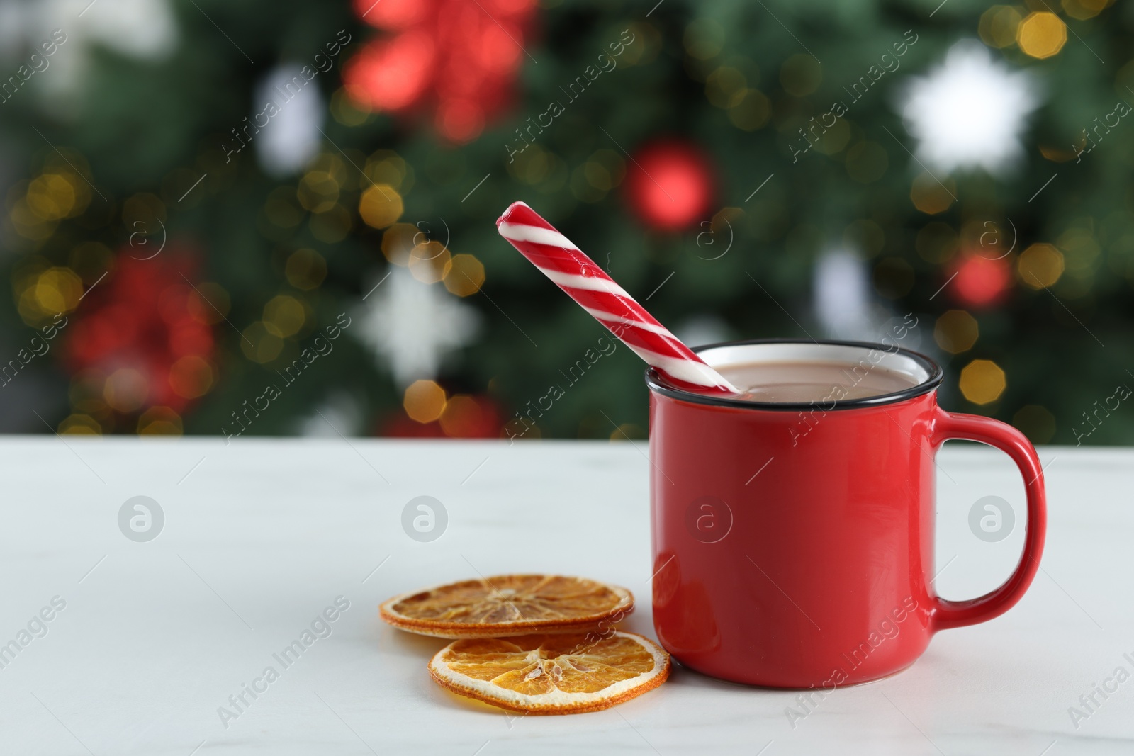 Photo of Tasty hot cocoa drink with candy cane in mug and dry orange slices on white table against blurred lights. Space for text