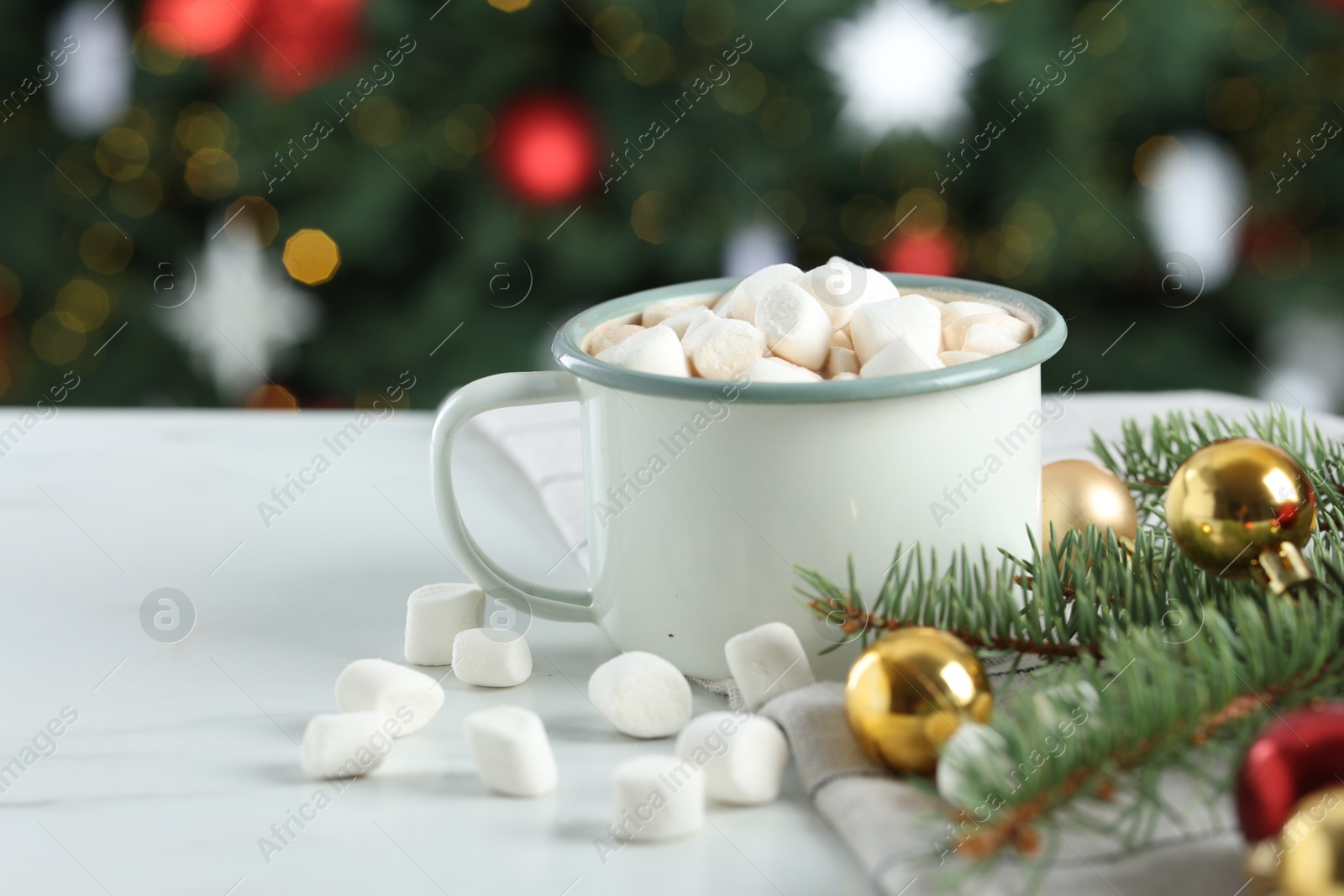 Photo of Tasty hot cocoa drink with marshmallows in mug and Christmas decor on white table against blurred lights, closeup. Space for text