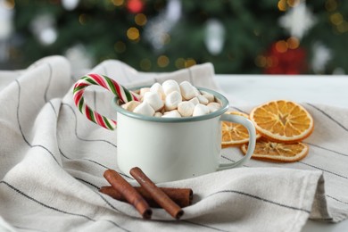 Photo of Tasty hot cocoa drink with marshmallows, candy cane in mug and spices on table, closeup