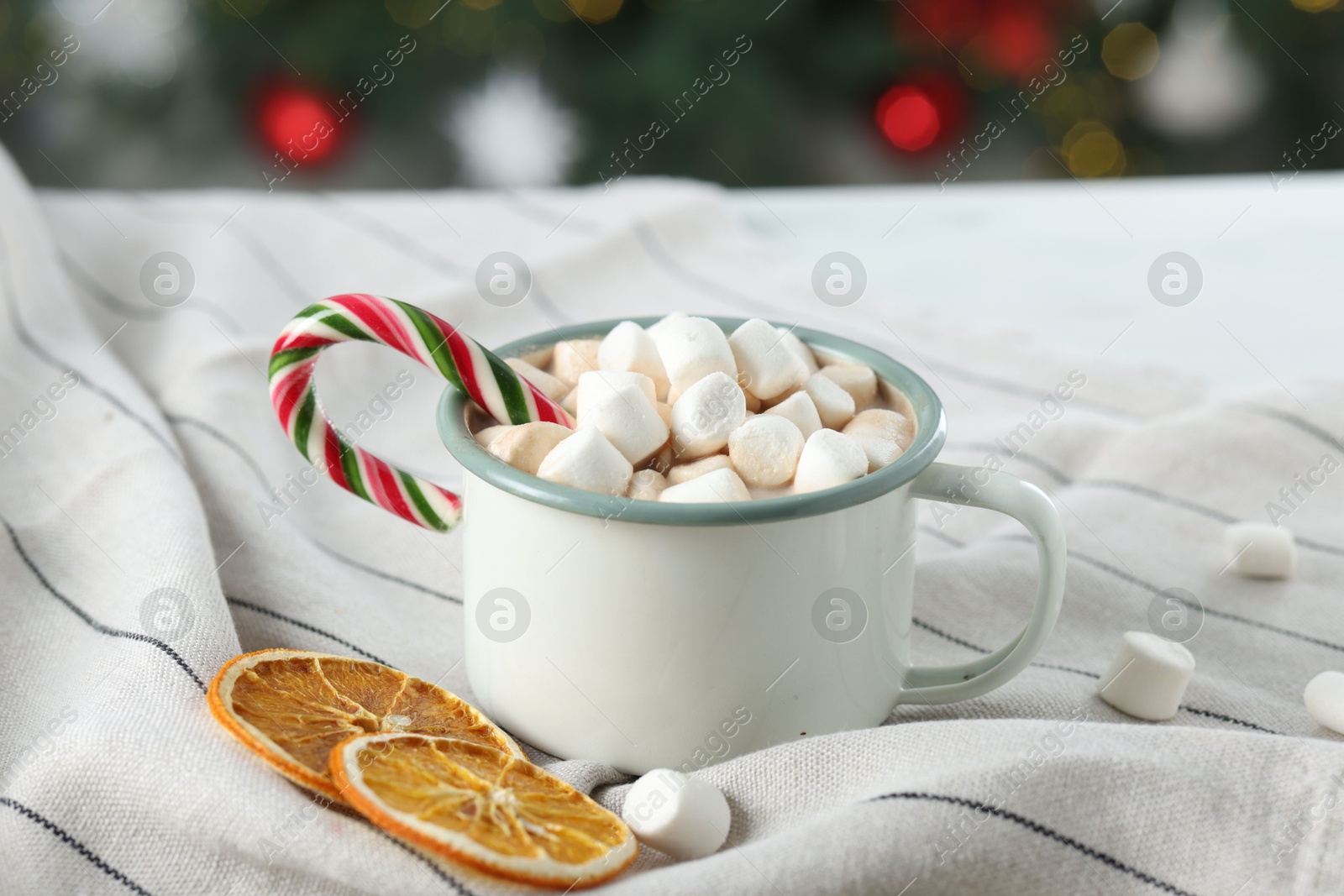 Photo of Tasty hot cocoa drink with marshmallows, candy cane in mug and dry orange slices on table, closeup