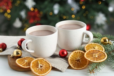 Photo of Tasty hot cocoa drinks in cups, fir branches, baubles and dry orange slices on white table against blurred lights, closeup