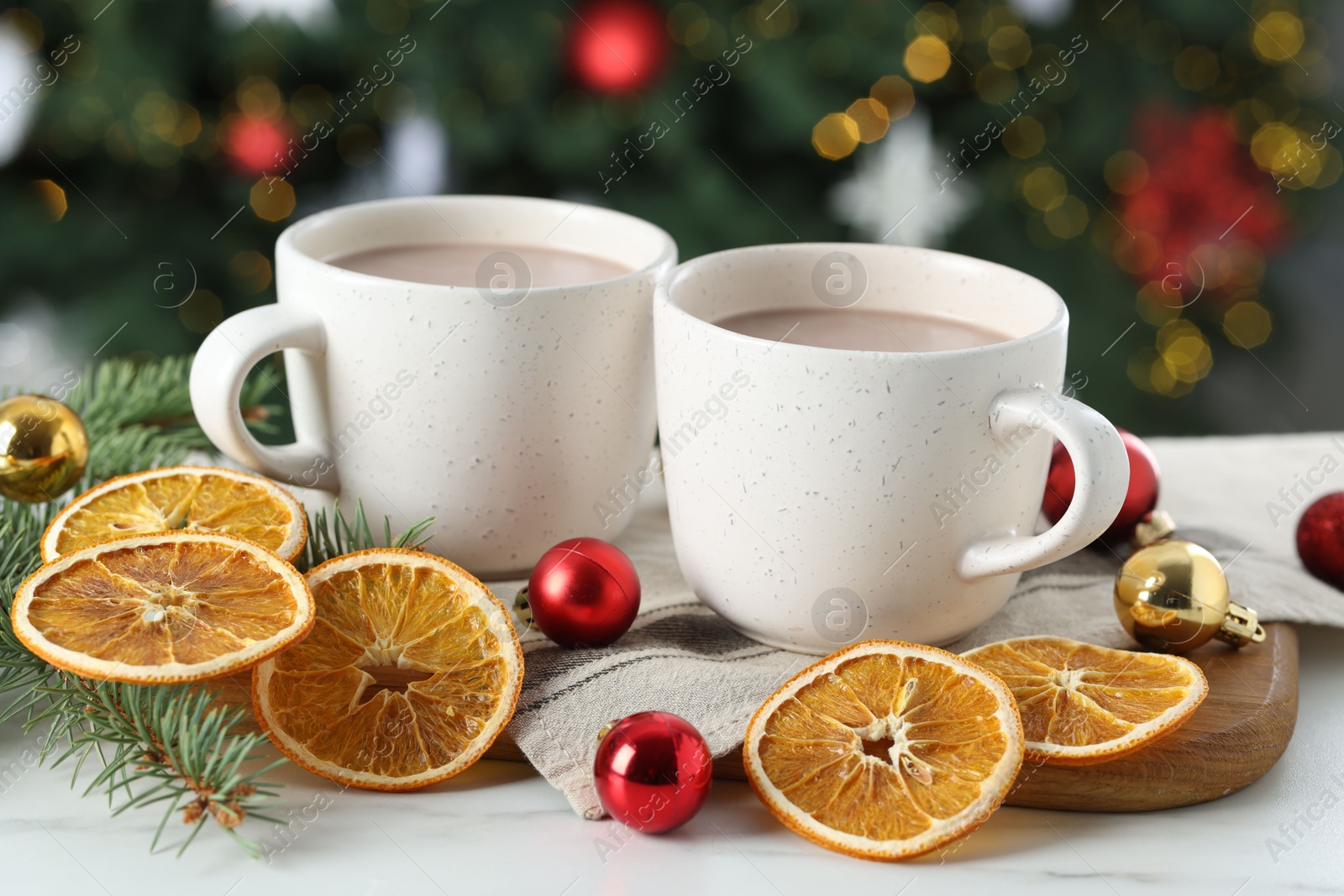 Photo of Tasty hot cocoa drinks in cups, fir branches, baubles and dry orange slices on white table against blurred lights, closeup