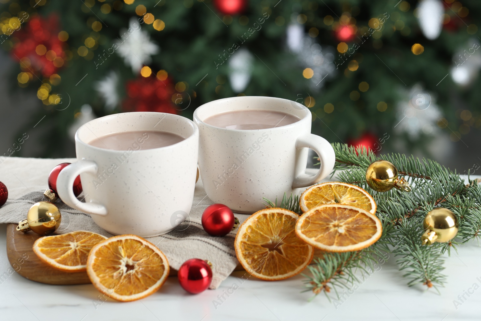 Photo of Tasty hot cocoa drinks in cups, fir branches, baubles and dry orange slices on white table against blurred lights