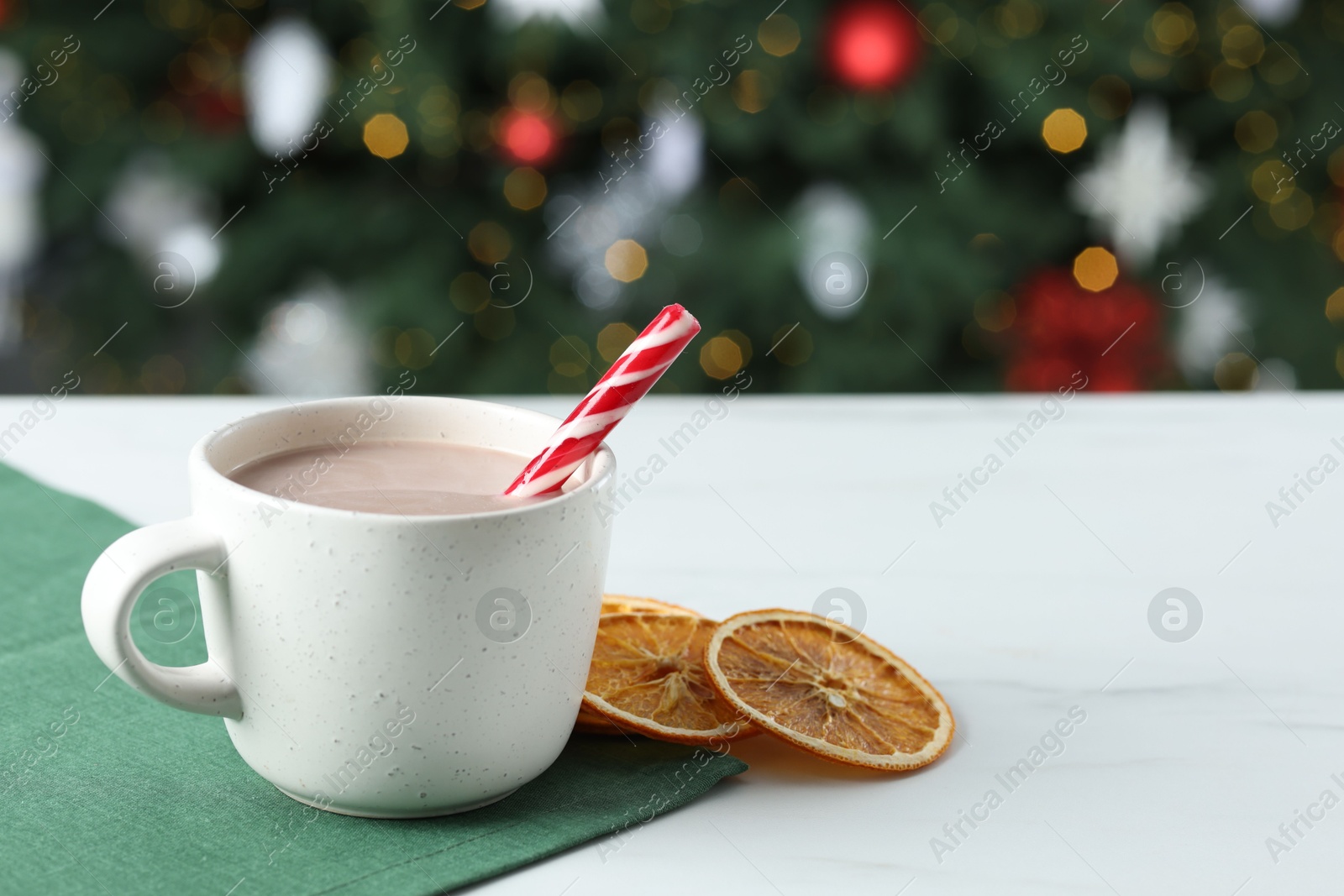 Photo of Tasty hot cocoa drink with candy cane in cup and dry orange slices on white table against blurred lights, closeup. Space for text