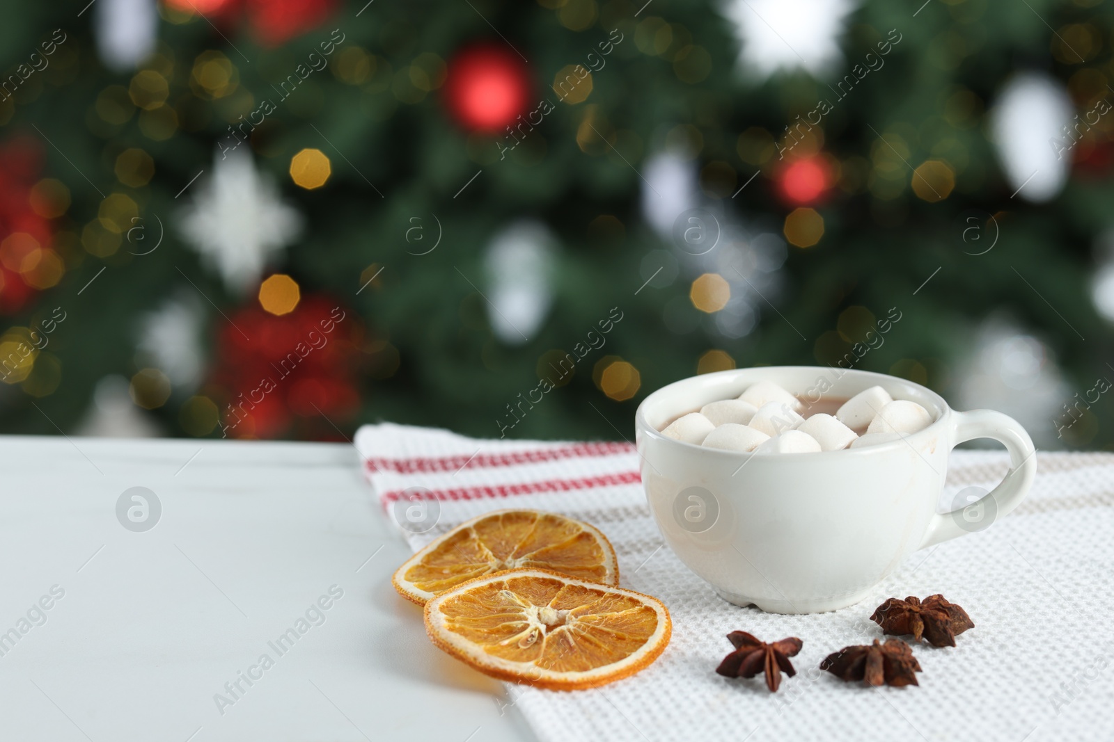 Photo of Tasty hot cocoa drink with marshmallows in cup, anise stars and dry orange slices on white table against blurred lights. Space for text