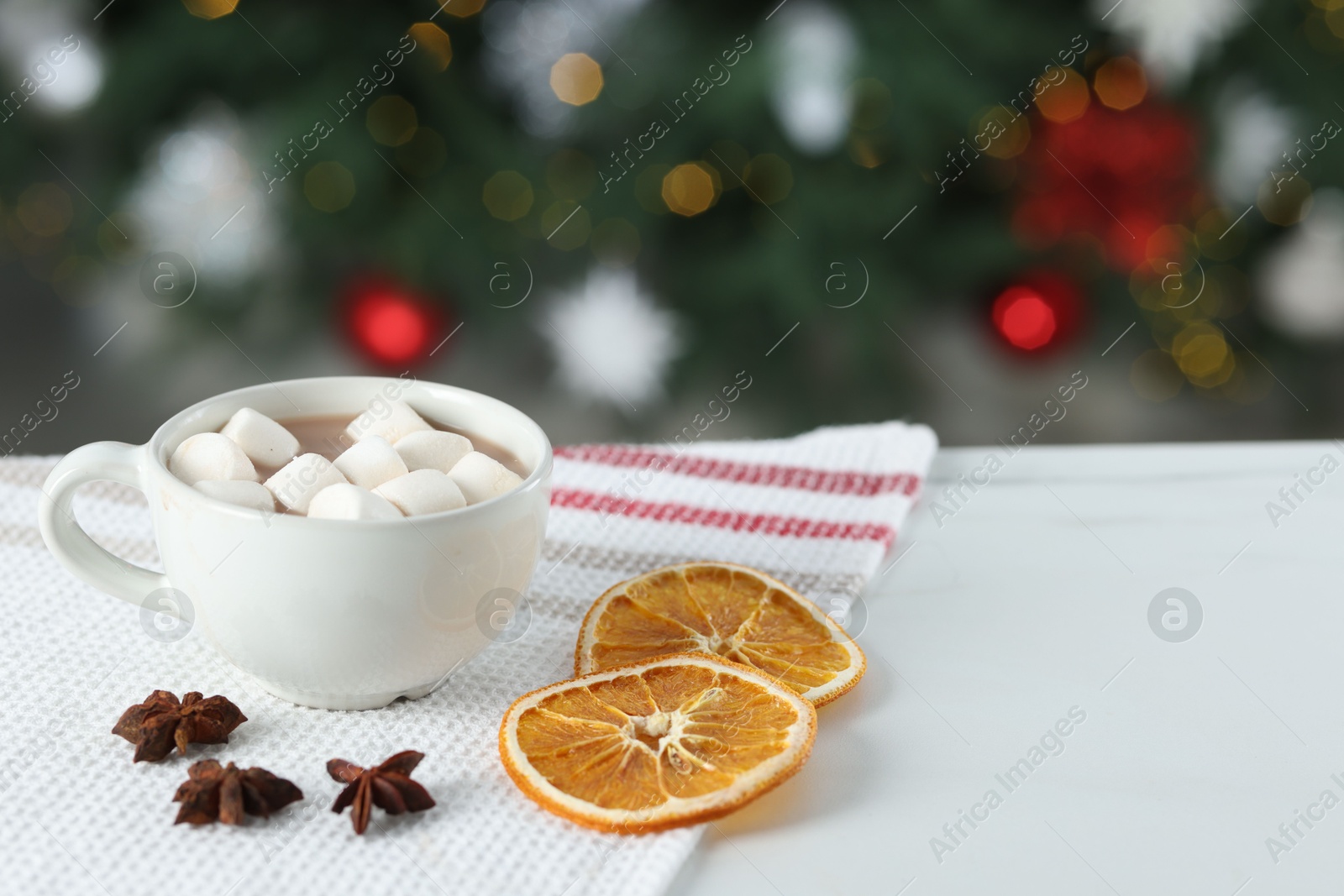 Photo of Tasty hot cocoa drink with marshmallows in cup, anise stars and dry orange slices on white table against blurred lights. Space for text