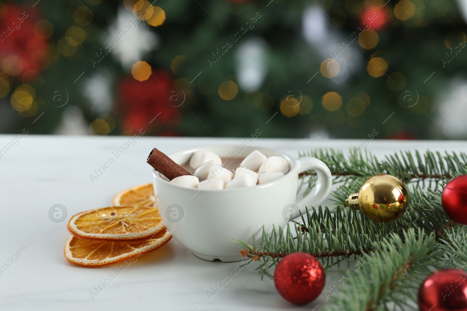 Photo of Tasty hot cocoa drink with marshmallows in cup, fir branches, baubles and dry orange slices on white table against blurred lights