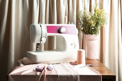 Photo of Sewing machine with fabric, flowers in vase and tools on chest of drawers indoors