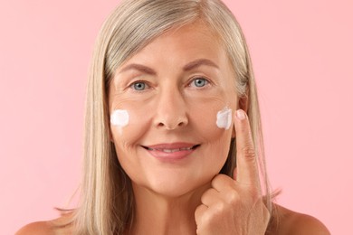 Senior woman applying face cream on pink background