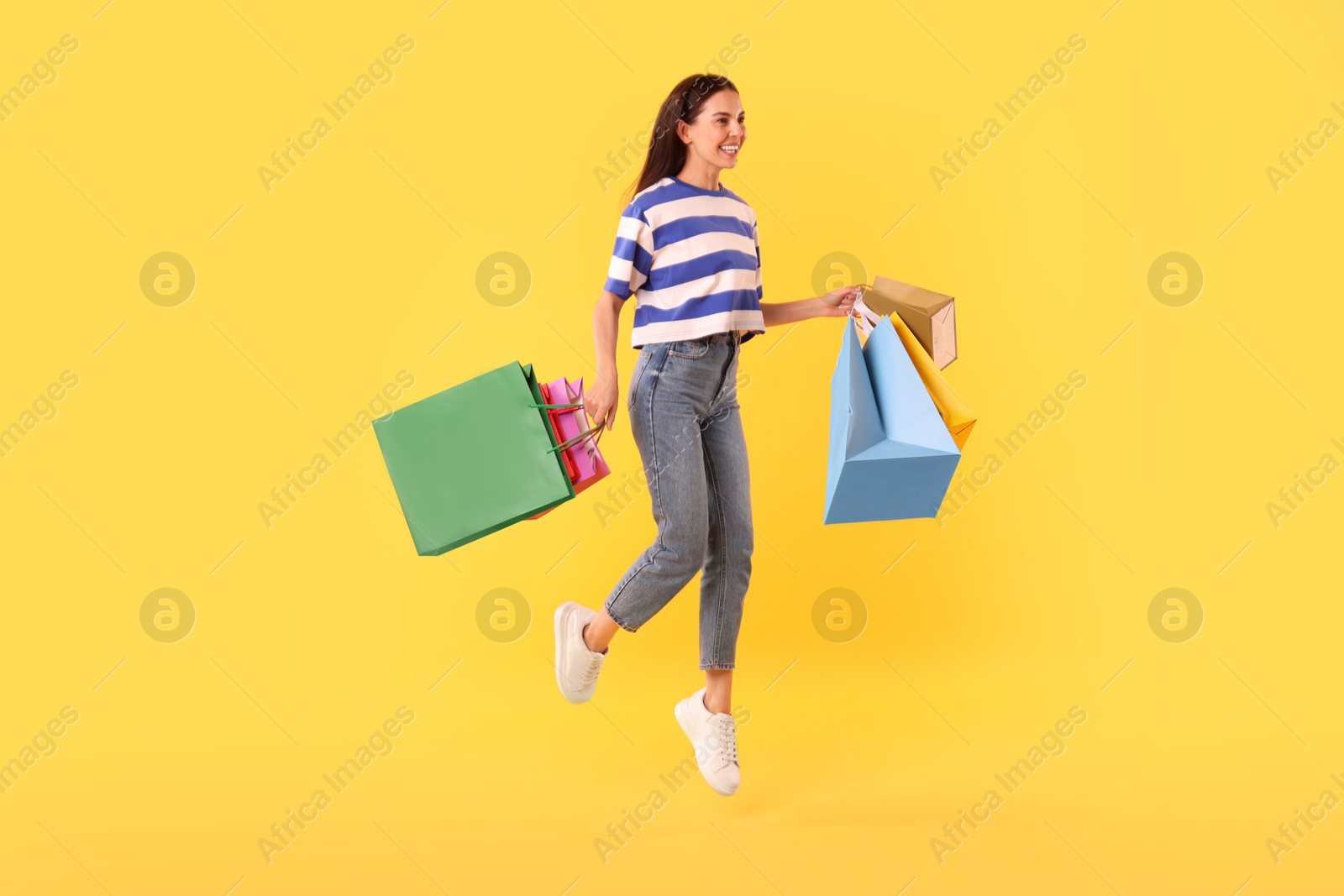 Photo of Smiling woman jumping with colorful shopping bags on yellow background