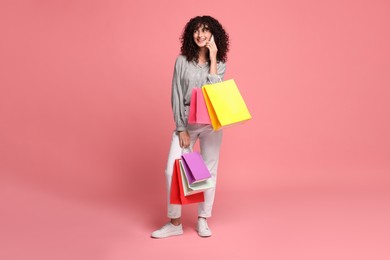 Photo of Smiling woman with colorful shopping bags talking by smartphone on pink background