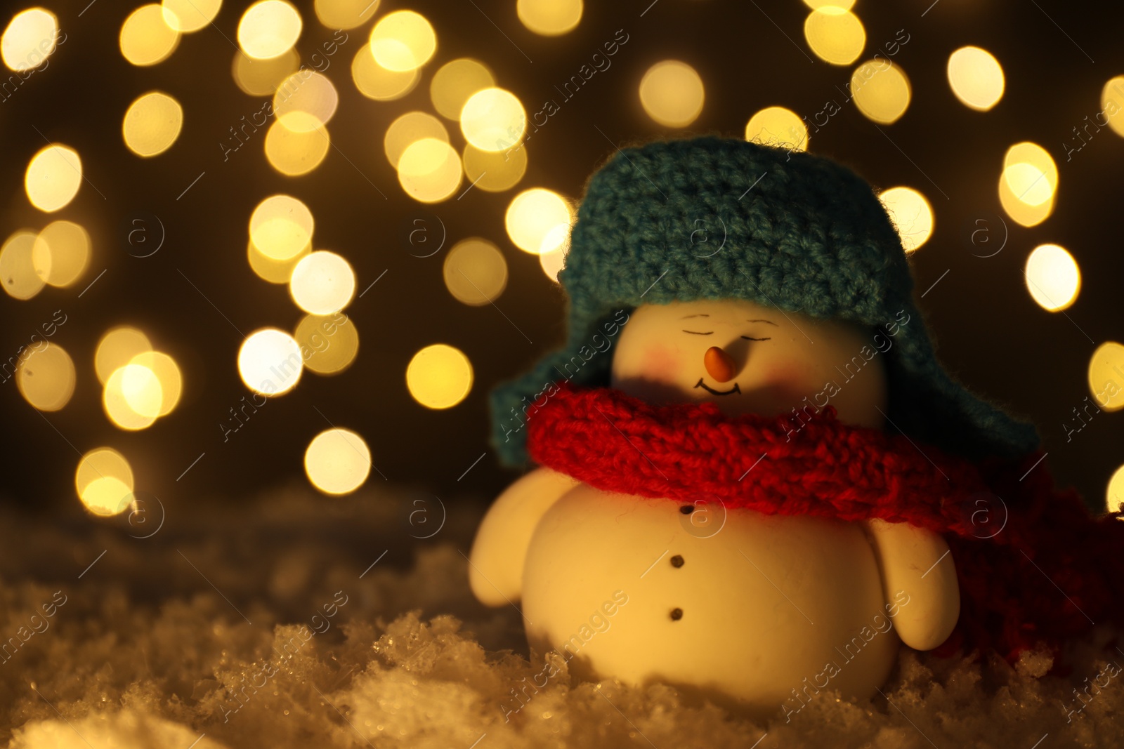 Photo of Cute decorative snowman on artificial snow against blurred lights, closeup. Space for text