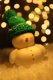 Cute decorative snowman on artificial snow against blurred lights, closeup