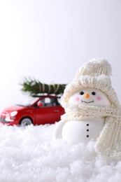 Photo of Cute decorative snowman and toy car with fir tree branches on artificial snow against light background, selective focus