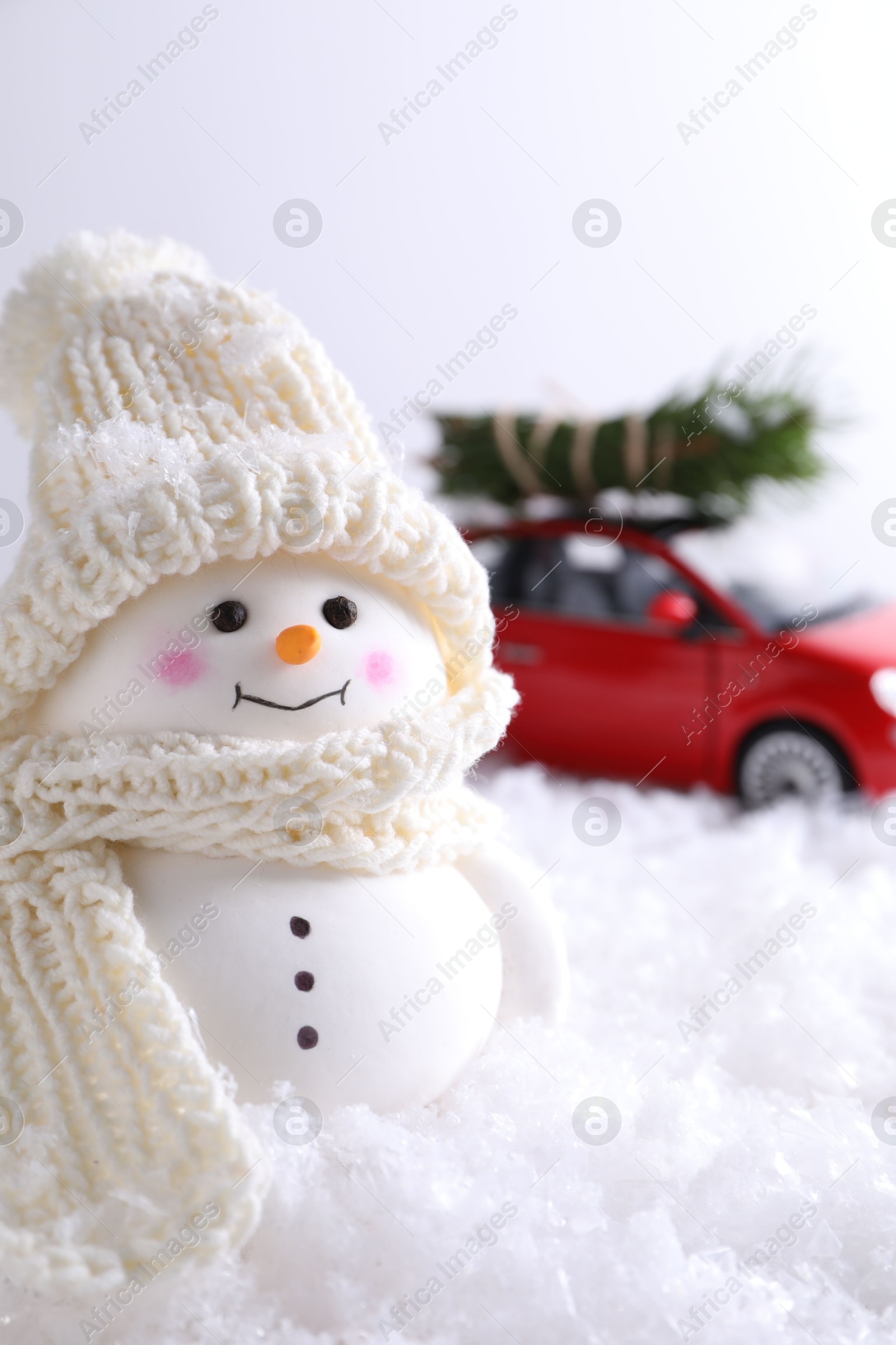 Photo of Cute decorative snowman and toy car with fir tree branches on artificial snow against light background, selective focus