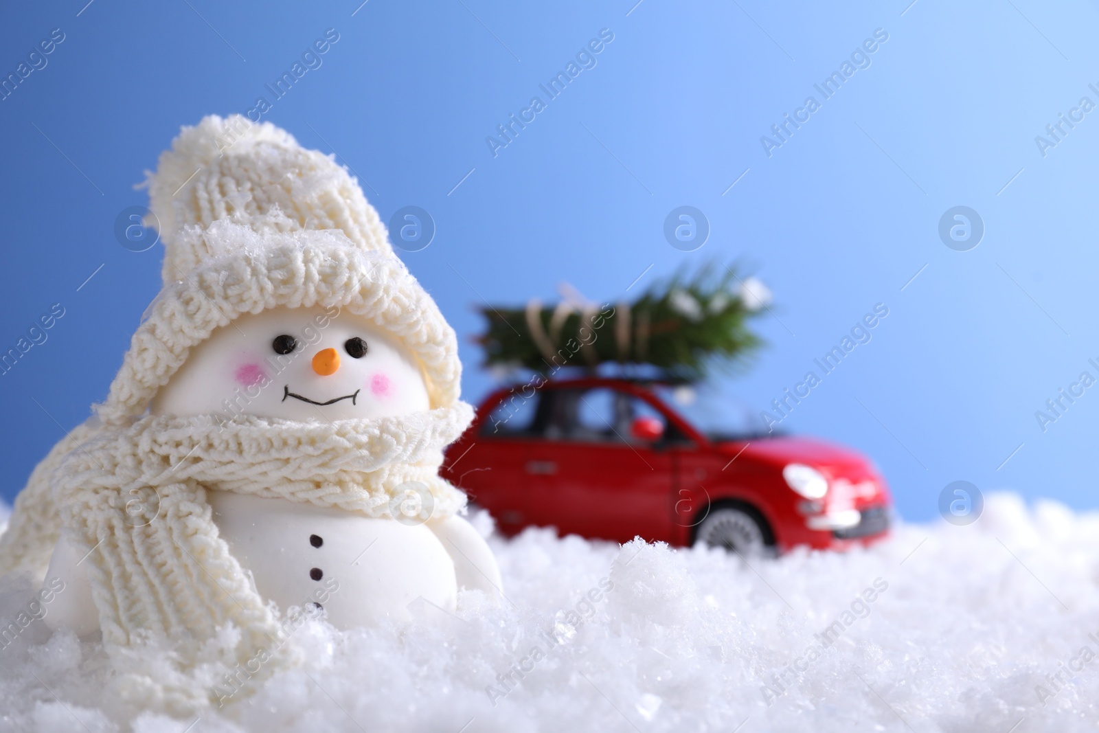 Photo of Cute decorative snowman and toy car with fir tree branches on artificial snow against light blue background, selective focus