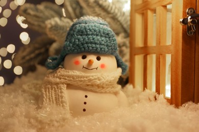 Photo of Cute decorative snowman and wooden lantern on artificial snow against blurred lights, closeup