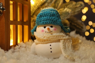 Photo of Cute decorative snowman and wooden lantern on artificial snow against blurred lights, closeup