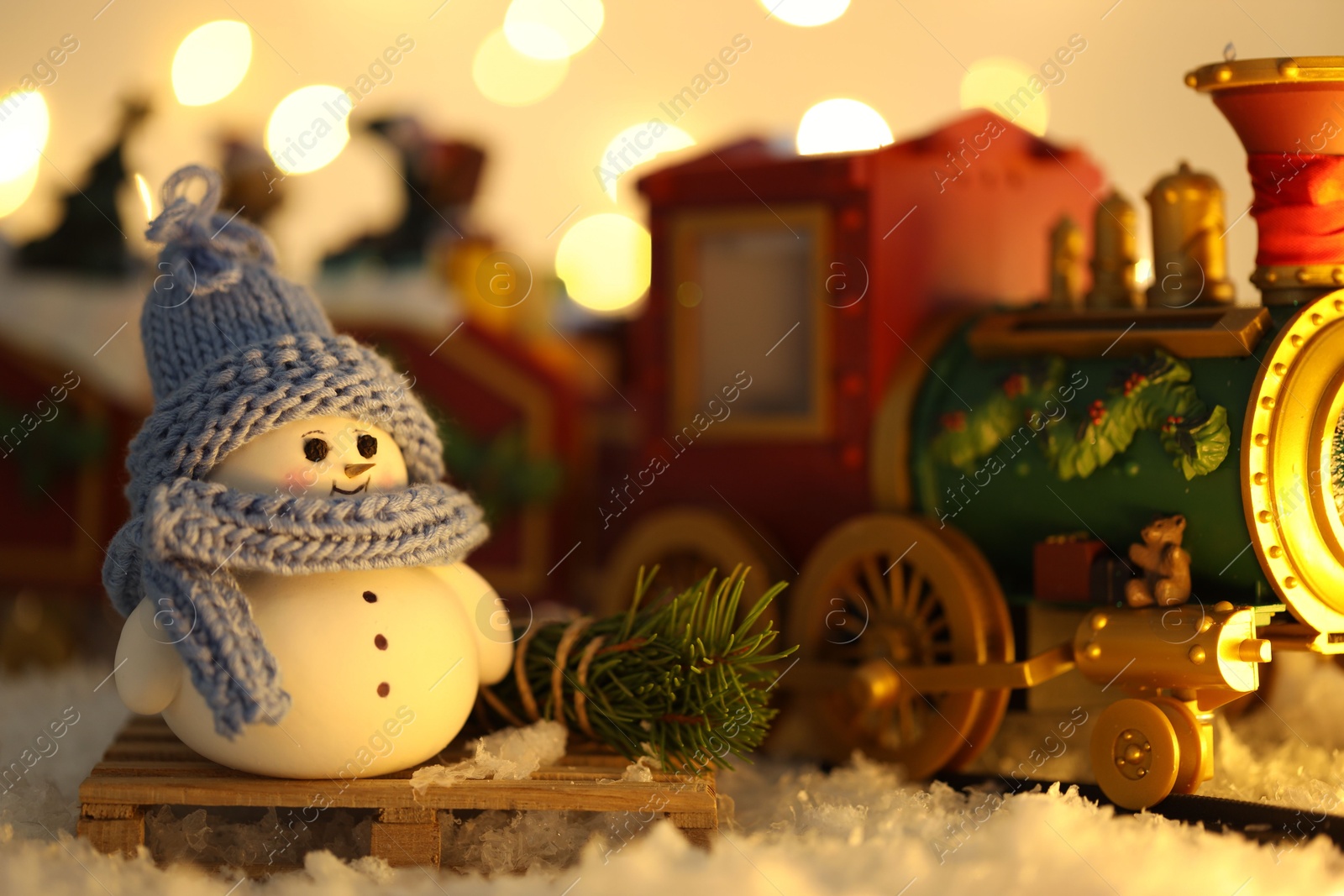 Photo of Cute decorative snowman and toy train on artificial snow against blurred lights, closeup