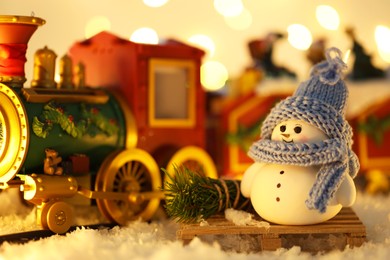 Photo of Cute decorative snowman and toy train on artificial snow against blurred lights, closeup