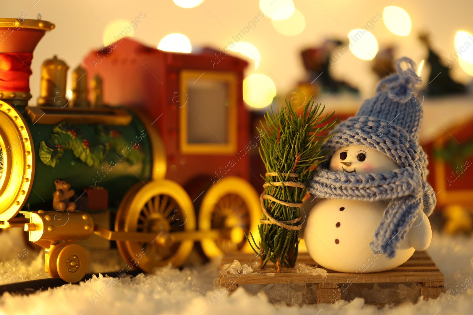 Photo of Cute decorative snowman and toy train on artificial snow against blurred lights, closeup