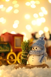 Photo of Cute decorative snowman, fir tree branches and toy train on artificial snow against blurred lights, selective focus