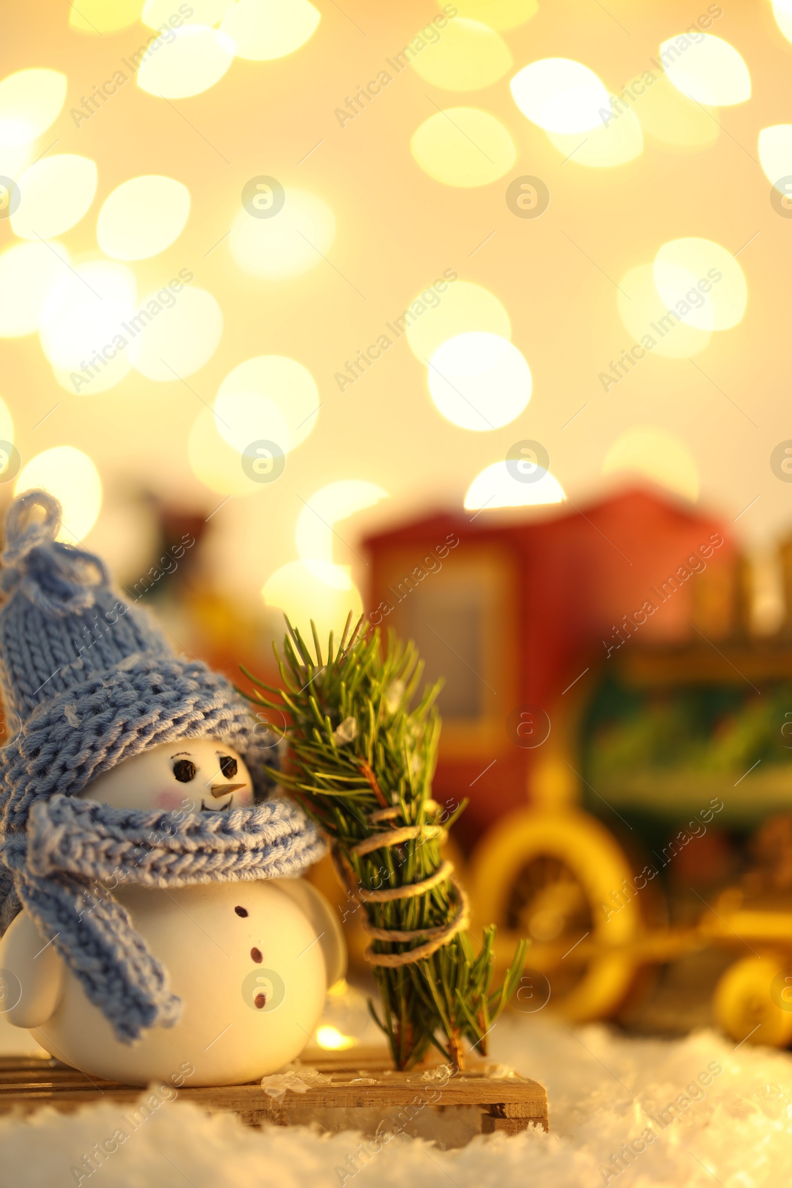 Photo of Cute decorative snowman, fir tree branches and toy train on artificial snow against blurred lights, selective focus