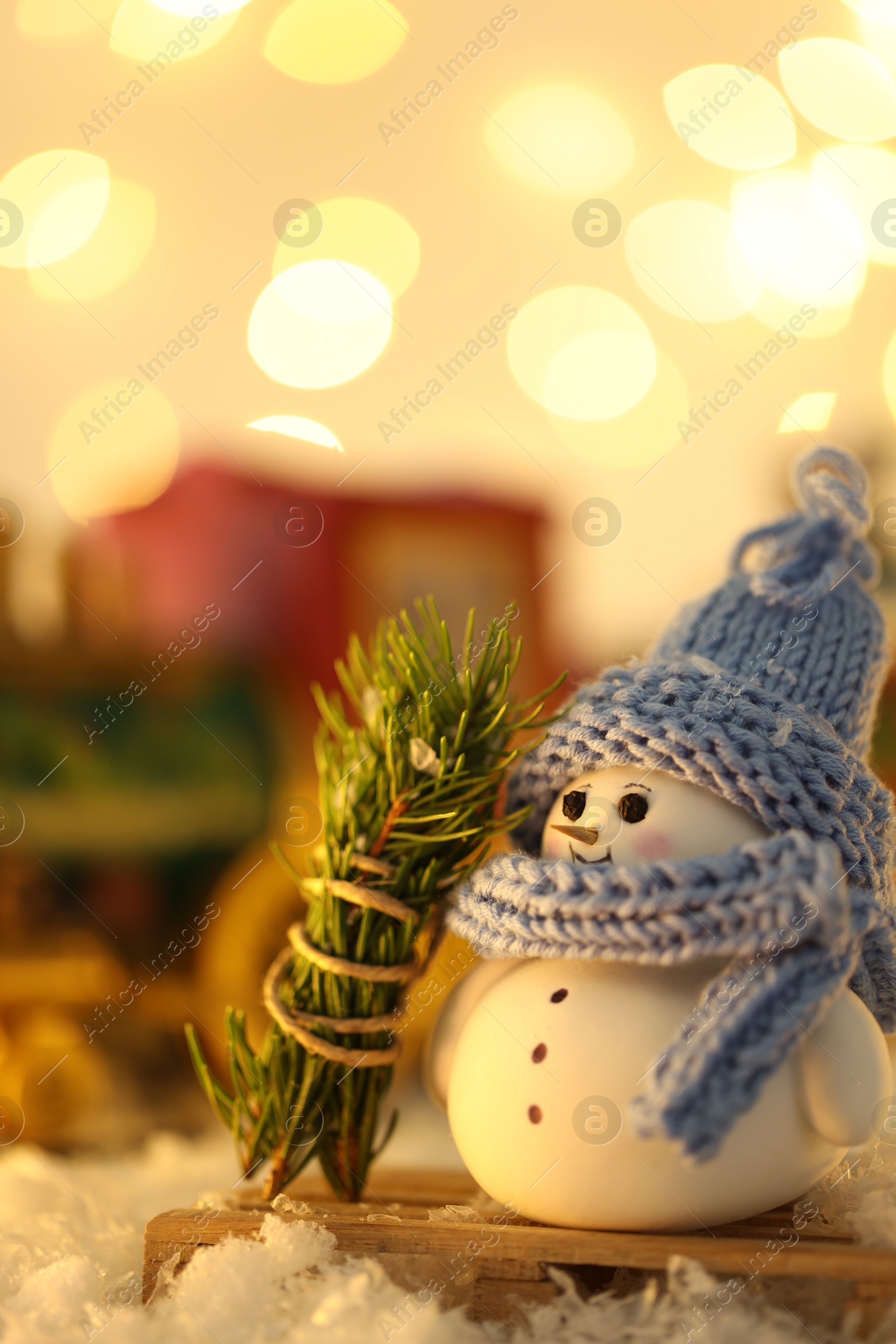 Photo of Cute decorative snowman and fir tree branches on artificial snow against blurred lights, closeup