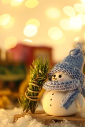 Cute decorative snowman and fir tree branches on artificial snow against blurred lights, closeup