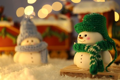 Photo of Cute decorative snowmen on artificial snow against blurred lights, selective focus