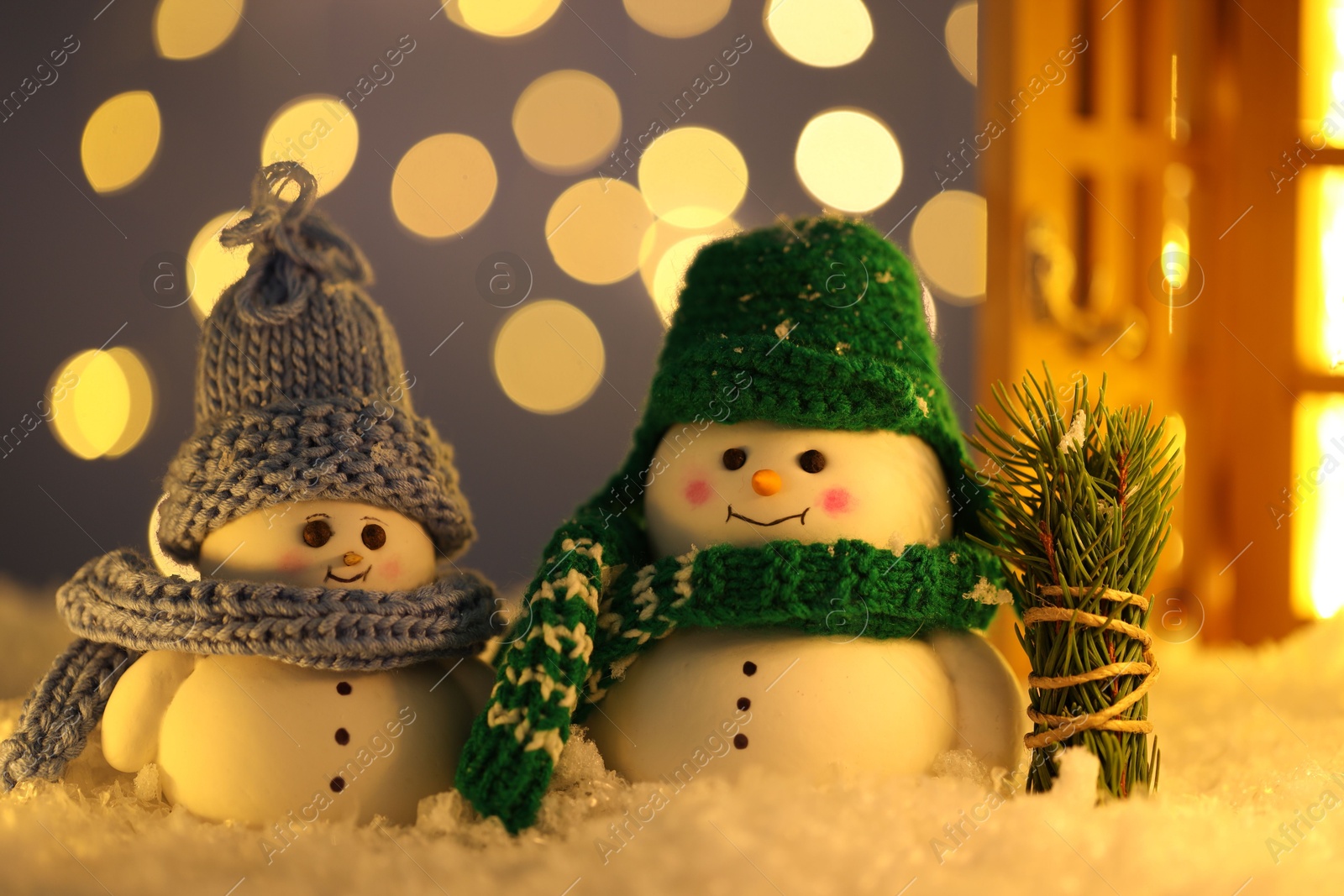 Photo of Cute decorative snowmen and fir tree branches on artificial snow against blurred lights, closeup