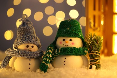 Photo of Cute decorative snowmen and fir tree branches on artificial snow against blurred lights, closeup